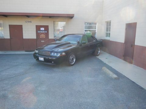 2003 Ford Crown Victoria - black full-size sedan with a raised cowl-induction hood parked in a lot beside a tan and maroon building.