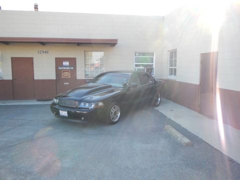 2003 Ford Crown Victoria - Front-quarter view of a lowered black full-size sedan parked beside a tan industrial building.