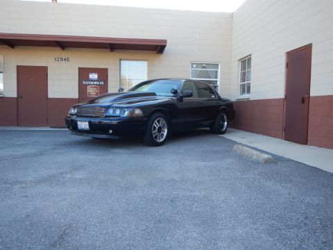 2003 Ford Crown Victoria - black full-size sedan angled in a parking lot beside a tan building with maroon trim.