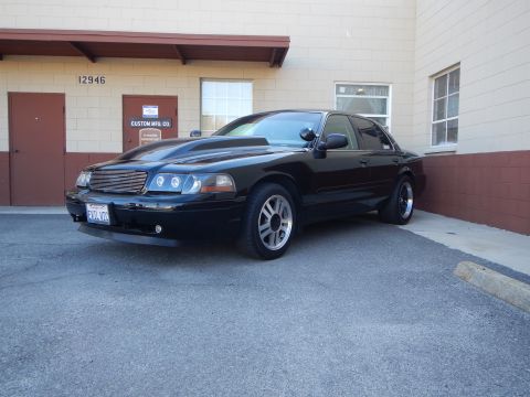 2003 Ford Crown Victoria - Front three-quarter view of a black full-size sedan with a chrome mesh grille, cowl hood and silver wheels.