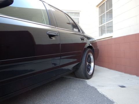 2003 Ford Crown Victoria - Low rear-quarter view of a black sedan fitted with a black wheel, parked beside a maroon-and-white wall.