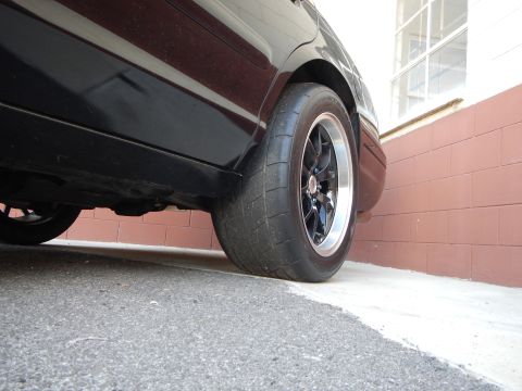 2003 Ford Crown Victoria - Ground-level rear-quarter shot of a black sedan with a black wheel and wide tire next to a maroon wall.