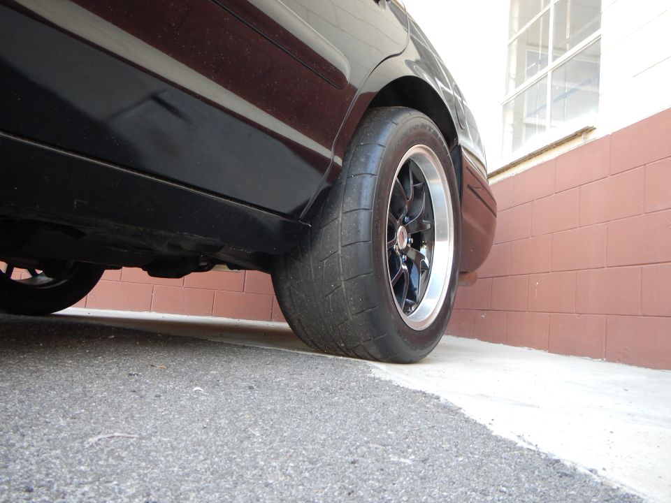 Low rear-quarter view of black sedan with black drag wheel and wide tire