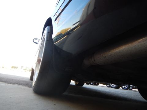 2003 Ford Crown Victoria - Low underside view of a black sedan's rear wheel showing a Ford mud flap, exhaust pipe and wide tire.