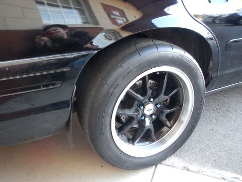 2003 Ford Crown Victoria - Black sedan's rear black wheel and tire, with the photographer's reflection visible in the glossy body.