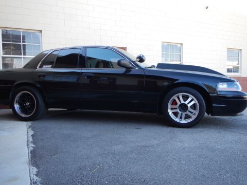 2003 Ford Crown Victoria - Side profile of a lowered black full-size sedan with black and silver wheels parked beside a white building.