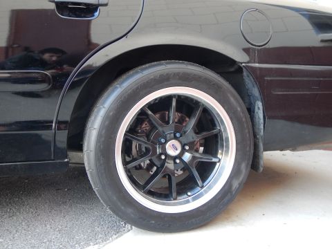 2003 Ford Crown Victoria - rear corner of a black car in a garage, fitted with a black machined-lip alloy wheel and a drag-style tire.