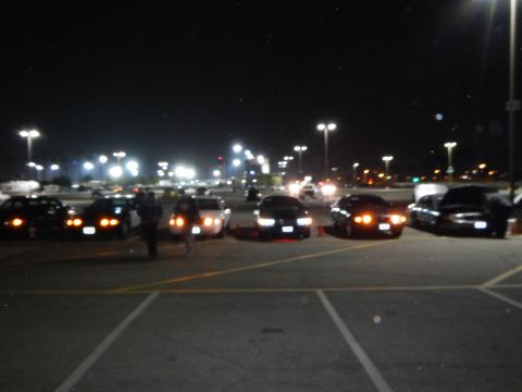 2003 Ford Crown Victoria - line of dark sedans parked side by side at night with amber parking lights lit, people standing nearby in a brightly lit lot.