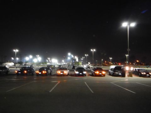 2003 Ford Crown Victoria - Several sedans lined up in a dark parking lot at night, headlights and parking lights glowing under stadium-style light poles.