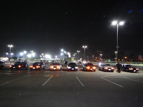 2003 Ford Crown Victoria - spread-out lineup of sedans in a night parking lot, lights lit, with a person standing beside the rightmost car.