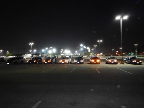 2003 Ford Crown Victoria - long row of sedans parked across a dark lot at night, their headlights and parking lights glowing beneath tall light poles.