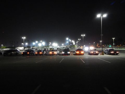 2003 Ford Crown Victoria - group of sedans parked at varied angles in a dark lot at night, lights lit, with a large illuminated parking area beyond.