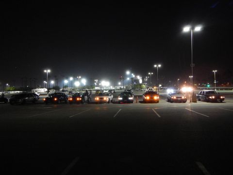 2003 Ford Crown Victoria - Numerous sedans arranged across a wide dark lot at night with parking and headlights glowing, distant lit buildings behind.