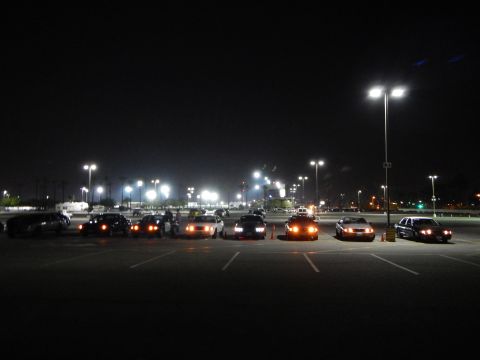 2003 Ford Crown Victoria - lineup of sedans with lights lit spread across a vast dark parking lot at night beneath distant stadium lighting.