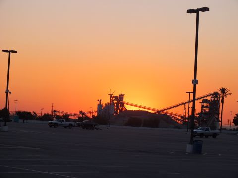 2003 Ford Crown Victoria - vivid orange sunset silhouettes industrial conveyor structures behind an empty parking lot with pickup trucks and light poles.