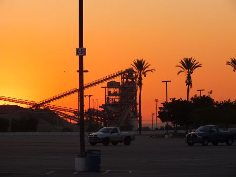 2003 Ford Crown Victoria - orange sunset silhouettes an industrial plant and palm trees, with two pickup trucks parked in the foreground lot.