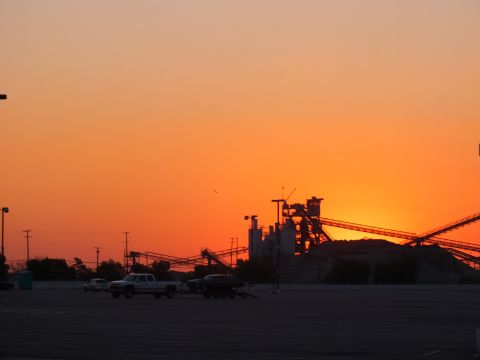 2003 Ford Crown Victoria - brilliant orange sunset behind silhouetted industrial conveyor towers, with a pickup truck and trailer in the empty lot.