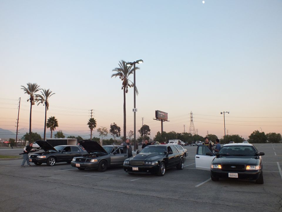 Sedans parked with hoods up and people gathered at dusk in a lot