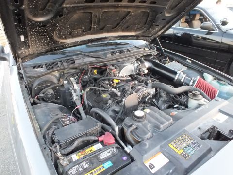 2003 Ford Crown Victoria - V8 engine compartment with a red cone air filter, a yellow battery, and wiring beneath the open hood of a sedan.