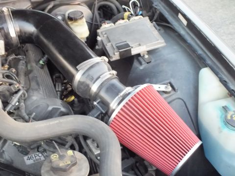 2003 Ford Crown Victoria - Close-up of a vehicle engine bay showing a red cone aftermarket air filter clamped to a black aftermarket intake tube near the brake fluid…