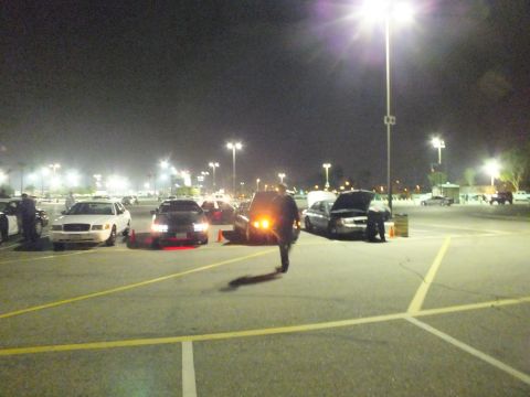 2003 Ford Crown Victoria - Nighttime car meet in a large lit parking lot with sedans parked in a row, one with its hood up, and a person walking across the lot.