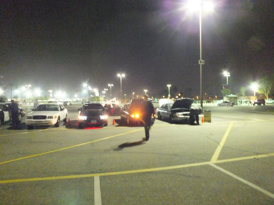 Nighttime parking lot with sedans lined up and a person walking, one hood open