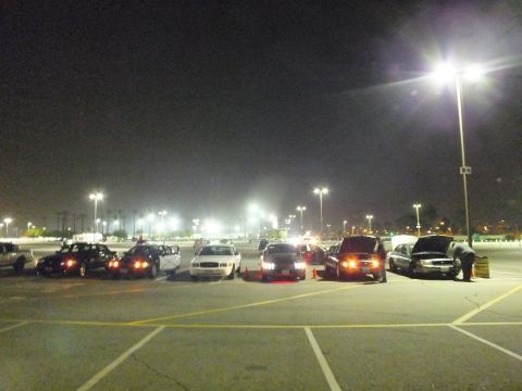 2003 Ford Crown Victoria - row of dark and white sedans parked at night in a large lot, with a person bent over an open hood at the far end.
