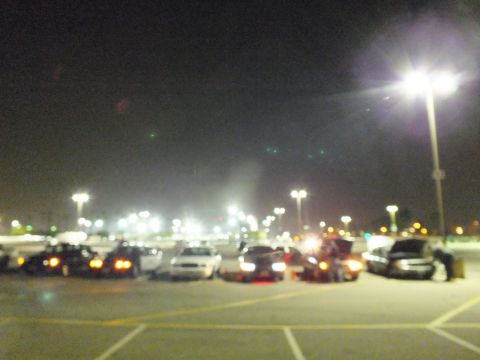 2003 Ford Crown Victoria - Blurry nighttime photo of multiple cars parked across a wide lot with bright overhead lights and tail lights glowing.