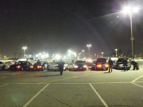 2003 Ford Crown Victoria - Nighttime gathering in a large parking lot with several sedans lined up, people standing around, and one car with its hood raised.