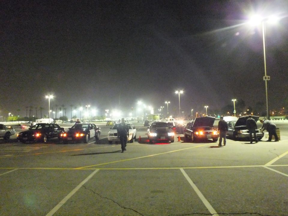 Cars and people gathered at night in a parking lot, one car with hood open