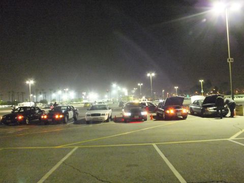 2003 Ford Crown Victoria - line of parked sedans at night with a person bent under an open hood at the right and traffic cones placed on the pavement.