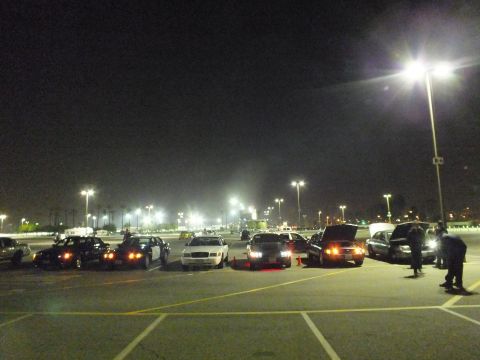 2003 Ford Crown Victoria - Wide nighttime lot scene with cars parked in a row, people standing on the right near an open hood, under bright overhead lights.
