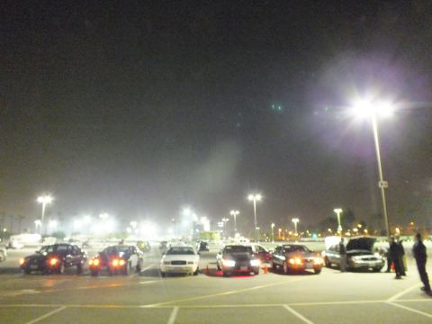 2003 Ford Crown Victoria - Dim wide-angle nighttime shot of a parking lot with rows of cars, traffic cones, and one car with its hood open at the right.