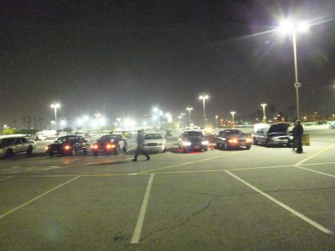 2003 Ford Crown Victoria - Nighttime parking lot with sedans parked at angles, one person crossing the lot and another standing by an open hood on the right.