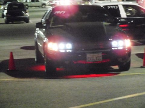 2003 Ford Crown Victoria - Front view of a lowered dark sedan with bright HID headlights and red underglow at night, positioned beside an orange traffic cone.