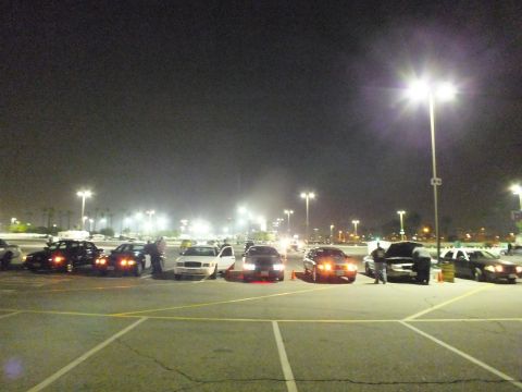 2003 Ford Crown Victoria - Wide nighttime view of a parking lot with several cars parked in a line, people standing among them, under tall light poles.