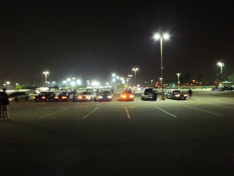 2003 Ford Crown Victoria - Distant nighttime view across a large parking lot showing cars parked in a row and people standing nearby under overhead lighting.