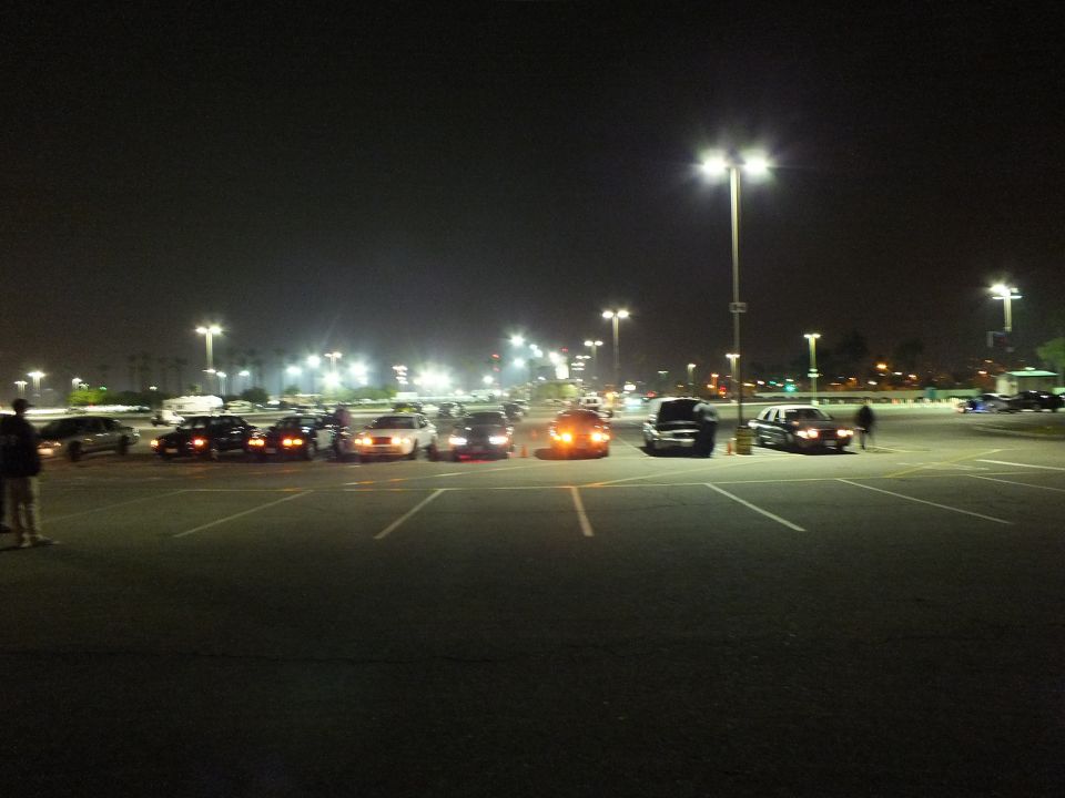 Distant nighttime view of cars parked in a large lit lot with people