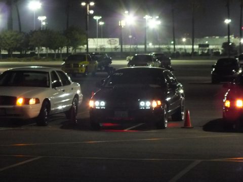 2003 Ford Crown Victoria - Nighttime lot scene with a dark sedan showing bright HID headlights beside a white sedan, with a yellow truck visible in the background.