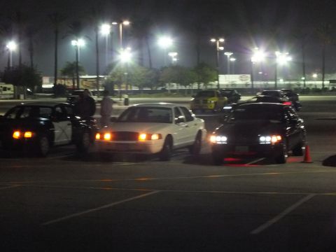 2003 Ford Crown Victoria - Nighttime parking lot scene with a white sedan in the center flanked by two dark sedans, headlights on, near a traffic cone.