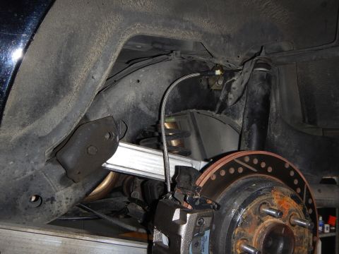 2003 Ford Crown Victoria - Underbody view of a rear suspension showing the coil spring, lower control arm, rubber bushings and a rusty vented brake rotor with caliper.