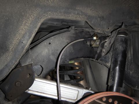 2003 Ford Crown Victoria - Underbody view of a rear suspension with the coil spring, lower control arm and a routed braided brake line near a yellow-zinc bracket.