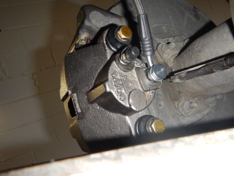 2003 Ford Crown Victoria - close-up of a cast brake caliper showing bleeder screw and gold banjo bolts, mounted near a concrete block wall.