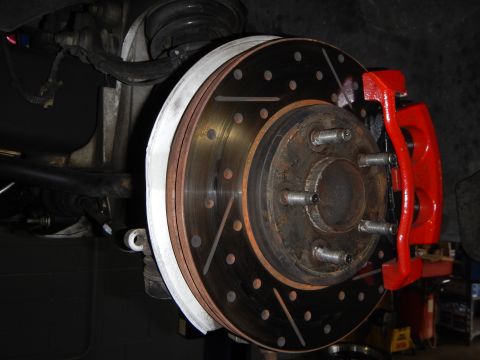2003 Ford Crown Victoria - angled underbody view of a slotted and drilled brake rotor with a red caliper and exposed lug studs.
