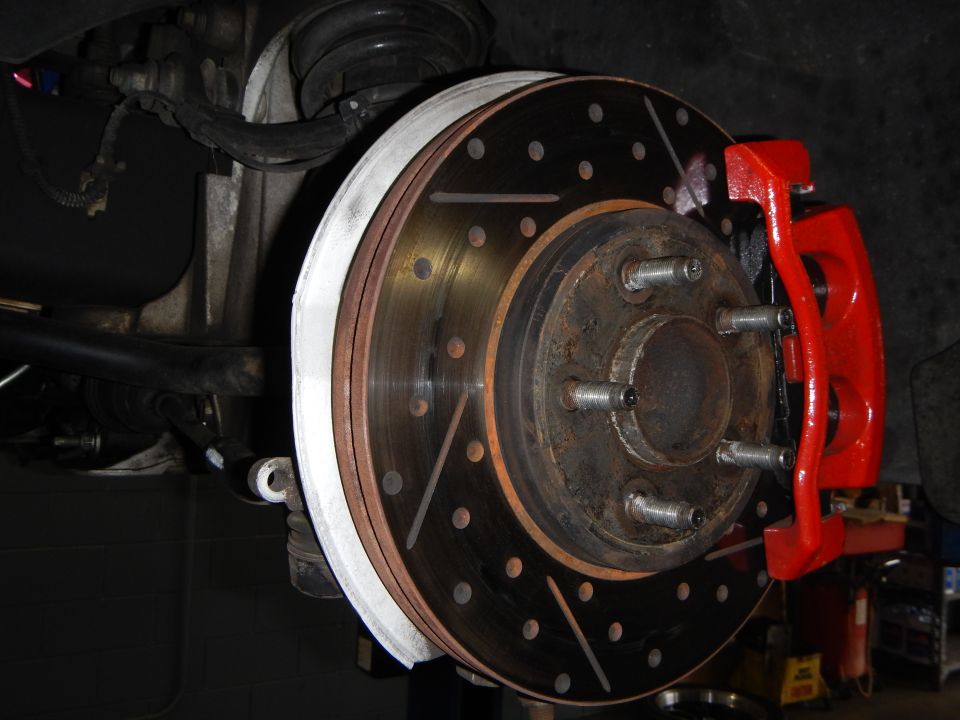 Slotted drilled brake rotor edge-on with red caliper and lug studs