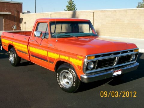 1979 Ford F-150 - Orange long-bed Ford F-Series pickup shot from the front three-quarter passenger side in a paved lot beside a tan block wall, wearing yello…