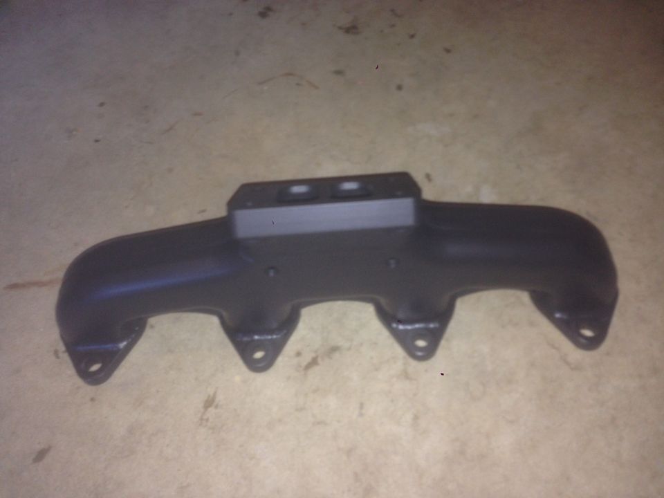 Black exhaust manifold with four ports and a central bracket on a concrete floor.