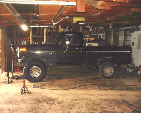 1985 Ford F-150 - A black 1985 Ford F-150 pickup truck is shown in a garage, with its front wheel elevated on a jack stand.