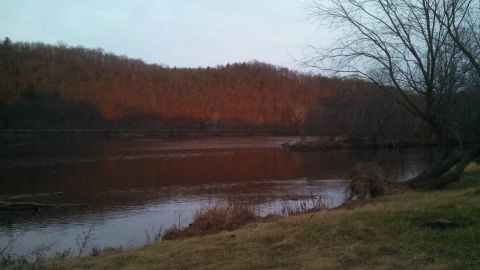 1979 Ford Bronco - A wide shot of a river flowing past a wooded hill at dusk. The setting sun casts a warm orange glow on the trees.
