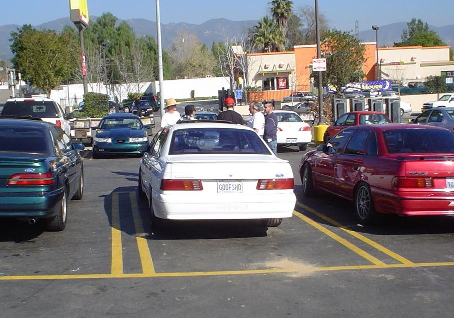 White 1993 Ford Taurus sedan with "GOOFSHO" license plate, parked in a lot with people around it.